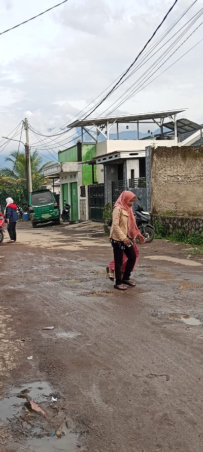 The image shows a street in Cangkuang, Indonesia, with two women walking on the muddy road. The women are wearing traditional Islamic clothing. One is wearing a pink headscarf and a long pink dress, the other is wearing a brown headscarf and a light brown jacket. The women are walking side by side, and the woman in the brown jacket is carrying a black bag. Behind them is a green van, parked on the side of the road. There is a small puddle in the middle of the road. The street is lined with buildings on both sides. There are a few trees on the left side of the image. There is a motorcycle parked near the right side of the image. There are power lines running above the street. The sky is cloudy and the sun is not visible. The location is near the coordinates latitude: -7.05, longitude: 107.54.