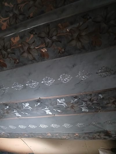 The image shows a ceiling decorated with a floral pattern. The pattern is divided into horizontal bands. The uppermost band features large, dark floral motifs with some lighter accents. Below this, there is a band with a marbled effect and a repeating pattern of smaller, intricate silver designs. The third band from the top also contains floral elements, but these are lighter in color, appearing as white or light grey outlines against a darker background. The lowest visible band mirrors the uppermost one, with dark floral designs. The lighting in the image is dim, and the perspective suggests the photo was taken from below, looking upwards at the ceiling. The environment appears to be an indoor space. There are no people visible in the image. The location is stated as Hlushky, Ukraine.