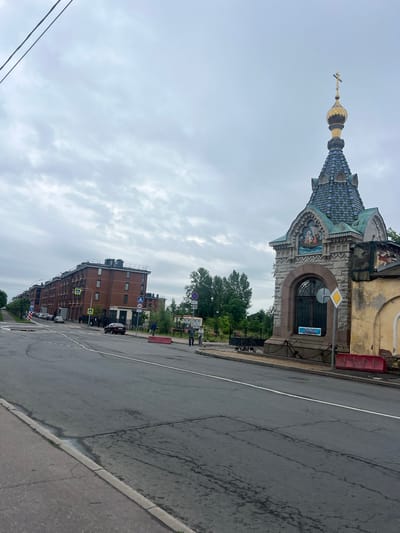 A medium-shot, eye-level, outdoor, slightly overcast day photograph captures a street scene in Kronstadt, Russia. On the right, a small, ornate stone building, possibly a chapel, features a multi-faceted roof tiled in shades of blue and green, topped with a golden onion dome and a cross. The building has an arched window with decorative metalwork, and above it, a mosaic depicts a scene with figures and a boat. A blue sign with white lettering hangs on the building. To the left of the chapel, a weathered yellow building abuts it. A low metal fence with thick chains separates the chapel from the sidewalk. In front of the chapel, a red concrete barrier is visible.

The street in the foreground is asphalt with visible cracks and white lane markings. A sidewalk lines the left side of the street. In the middle ground, several people are walking on the sidewalk and crossing the street. On the far left, a row of modern, multi-story brick apartment buildings stretches into the distance. Trees and greenery are visible behind the buildings and to the right of the chapel. Several cars are parked along the street and on the sidewalk. Traffic signs, including speed limit signs, pedestrian crossing signs, and parking restrictions, are posted along the road. The sky is cloudy and muted.