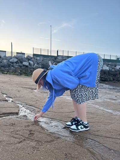 A person wearing a blue hooded sweatshirt, a short black and white floral dress, and black and white Nike Dunk sneakers stands on a sandy beach with water running over the sand. The person is bending over, reaching out with their right hand to touch the water. They are wearing a straw hat with a black band. Behind them is a seawall made of large rocks. The sky is light blue. The location appears to be a beach near a seawall in Gijón/Xixón, Spain.