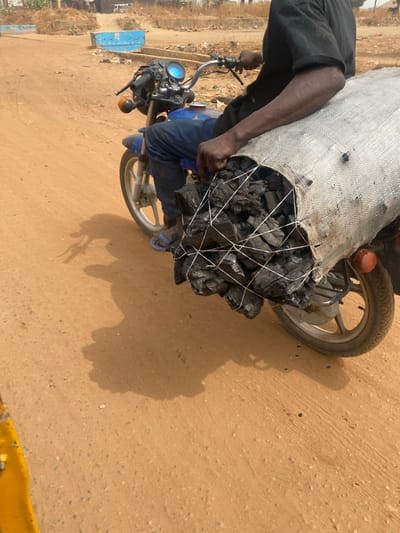 A man rides a motorcycle laden with a large sack of charcoal. The charcoal is bound by white netting, overflowing from the bag onto the motorcycle's front. The rider wears dark clothing and blue sandals, his dark skin contrasting with the bright tan dirt road. The bike's shadow stretches across the ground, suggesting a sunny day.