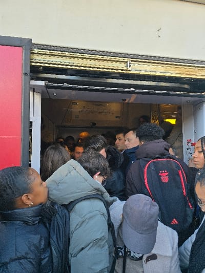 The image captures a densely packed public transport vehicle, likely a train or tram, viewed from an exterior perspective through an open door. A large group of people, appearing mostly young adults or teenagers, fills the interior, standing shoulder-to-shoulder with little to no visible space between them. Their faces are largely obscured or partially visible due to the extreme crowding.
Many individuals are wearing heavy winter coats or jackets in shades of black, grey, and dark green, suggesting cool or cold weather. Several backpacks are visible, including a prominent black Adidas backpack with red accents and a white logo on it. One person wears a grey baseball cap. A person in the foreground has a small gold earring visible.
The scene depicts a busy commute during daytime, evidenced by the bright natural light illuminating the interior of the vehicle and the exterior platform area. The transport vehicle appears to be stationary with its doors open. On a white panel inside the vehicle, some faint, unreadable text or diagrams are visible. A red panel or sign is partially seen in the immediate foreground, to the left of the open door. The overall impression is one of congestion typical of peak travel hours in an urban environment like Nice, France, as indicated by the context. No specific emotions are clearly discernible from the obscured faces.