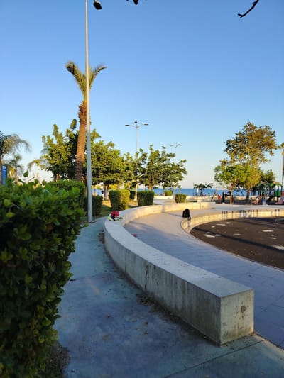 The image shows a paved public area with a curved concrete wall in the foreground. To the left, a dense green hedge partially obscures a concrete walkway. Behind the wall, a dark asphalt surface is visible, possibly part of a playground or recreational area, with some scattered leaves. In the middle ground, a palm tree stands tall next to other leafy green trees. A lamppost is situated between the palm tree and the hedge. Further back, the sea is visible with people walking along the shore and a playground with colorful structures. The sky is clear and blue. The scene suggests a public park or promenade in a coastal city, likely in a warm climate. Based on the vegetation and architecture, this scene is consistent with the coastal areas of Antalya, Turkey, possibly in a park like the one along Konyaaltı Beach or near the Old Town (Kaleiçi) with its sea views.