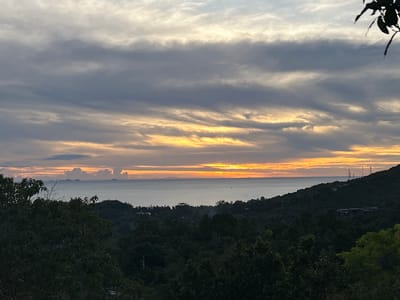 The image shows a sunset over the sea, likely taken from an elevated position in the Baan Hin Kong area of Thailand. The sky is filled with layers of clouds, colored with hues of grey, yellow, and orange from the setting sun. Below the horizon is the calm sea, with some islands visible in the distance on the left. The foreground is dominated by lush, dark green vegetation, suggesting a dense forest or jungle environment. On the right are the trees and some buildings with tall cellular towers.