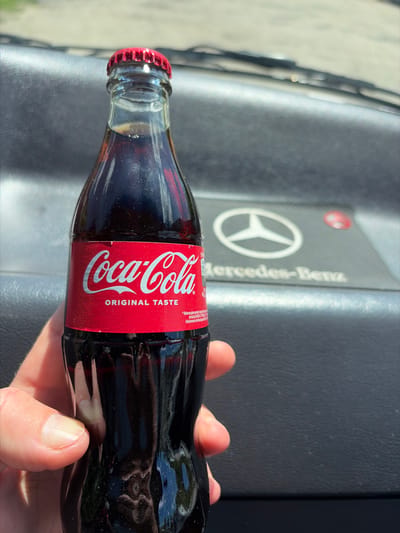 A person's hand holds a glass bottle of Coca-Cola with a red label and a red cap. The bottle appears to be lying on the dashboard of a car, specifically a Mercedes-Benz, as indicated by the logo visible on the dashboard. The background is slightly blurry, showing what appears to be an outdoor environment. It is not possible to determine the location of Wisła, Poland, from this image.