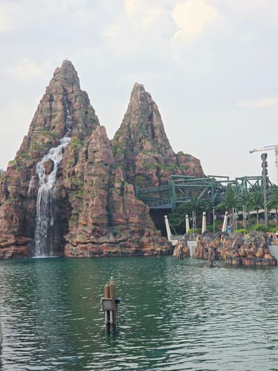 This is a photograph of a man-made rock formation with a waterfall, situated next to a body of water. The rock formation appears to be part of a theme park or attraction, as evidenced by the presence of roller coaster tracks and supports integrated into the structure, along with palm trees and other greenery. In the water, a small floating platform with some equipment is visible. The sky is overcast, suggesting it may be a cloudy day. The scene is brightly lit, indicating it is daytime. The rock formation is tall and imposing, with various textures and colors suggesting weathered stone. Water cascades down from multiple points on the rocks, creating a dynamic visual element. The overall impression is one of an engineered, fantastical landscape. There is no visible text in the image. The location context provided is Tongzhou District, China.