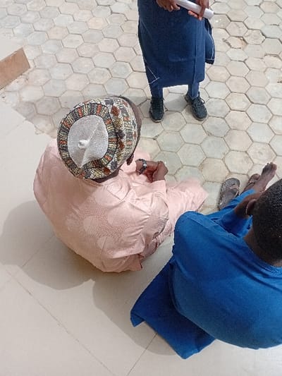 The scene is an outdoor area in Kwazailewa, Nigeria, under direct sunlight, evidenced by sharp shadows. Three individuals are present. In the mid-ground, a person is seated with their back to the viewer, wearing a light pink, dotted traditional garment and a patterned kufi cap with a light grey central dome. This individual is holding a small object in their hands, and a ring is visible on one finger. In the immediate foreground, a second individual is partially visible from the back, seated and wearing blue attire; a pair of grey sandals are near their feet. A third person stands in the background, visible from the waist down, wearing dark blue trousers and black athletic shoes, holding a rolled white document with both hands. The ground beneath the seated individuals is a light-colored, smooth tiled surface, which transitions to light grey hexagonal pavers in the background where the standing individual is positioned.