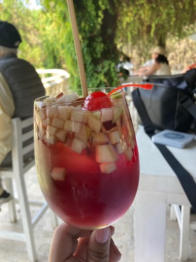The image depicts a glass of fruit cocktail held by a hand. The cocktail contains diced fruit and a maraschino cherry on a toothpick. The background shows a park-like setting with trees, white patio furniture and a black backpack. The location may be Tehuixtla, Mexico, as per the prompt.