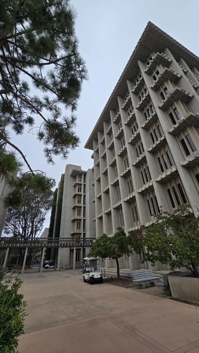 The image shows the modernist architecture of the UCSD Geisel Library in La Jolla, San Diego, California. The building, constructed from precast concrete, features a distinctive brutalist design with repetitive fenestration and cantilevered elements. In the foreground, a paved walkway leads towards the library. A white golf cart is parked on the right side of the walkway, near a concrete bench and some trees. To the left, a walkway is covered by a wooden structure. Pine trees with their needles are visible in the upper left corner, partially obscuring the sky, which is overcast. Other buildings with similar concrete construction and staggered window patterns are visible in the background.
