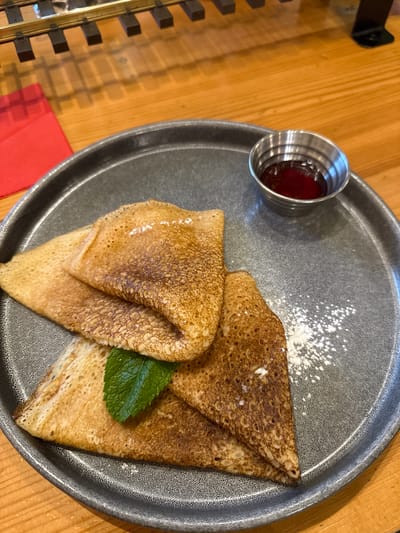 A close-up shot captures a plate of crepes on a wooden table. The plate, a dark gray, textured circle, holds three folded crepes, two layered on top of one, with a small bunch of fresh mint leaves tucked between them. A small silver metal ramekin filled with a dark red jam or sauce sits to the upper right of the crepes. Powdered sugar is lightly sprinkled on the plate. In the background, a red napkin is visible to the left, and above that, what appears to be a model train track is situated on the wooden surface. The lighting suggests an indoor setting. The image is devoid of people, actions, or interactions, focusing solely on the presentation of the food and its immediate surroundings.
