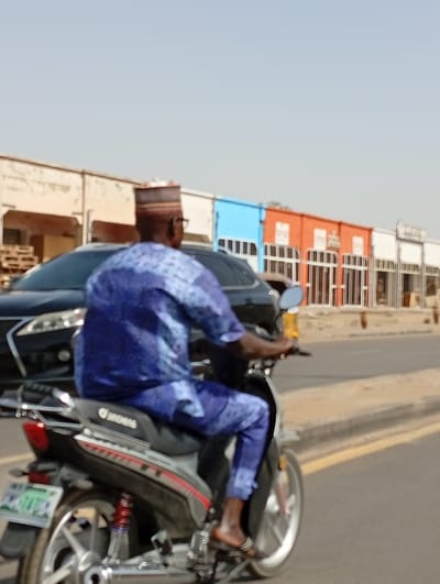 The image depicts an outdoor street scene in Sabon Gari West, Nigeria, under a clear, bright sky. The primary subject is a male individual operating a JINCHENG brand motorcycle, moving away from the camera's perspective. He is dressed in a blue and white patterned traditional long-sleeved tunic and matching trousers, paired with a multi-colored patterned cap and dark-framed eyeglasses. His feet are clad in dark sandals. The motorcycle is silver and black, featuring a visible rear wheel with a red-springed shock absorber and a blurred green license plate displaying white text. A dark-colored sedan is partially visible behind and to the left of the motorcycle. In the background, a row of single-story commercial buildings lines the street. These structures are painted in varying colors, including off-white, light blue, and orange, each featuring multiple window or door openings equipped with security bars. The road surface consists of asphalt, with a yellow painted curb line visible on the right side of the frame.