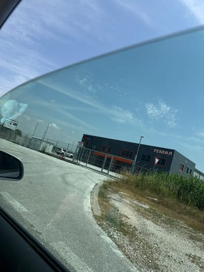 The image depicts a daytime scene in Dragor, Bulgaria, viewed from inside a vehicle through a tinted side window. The upper left portion of the image is obstructed by the vehicle's interior. A blue sky with scattered wispy clouds is visible above.
In the foreground, an asphalt road curves to the left, bordered by a concrete curb and a gravel shoulder. Dry grass and taller green reeds line the right side of the road.
Beyond the road and shoulder, a chain-link fence encloses a commercial or industrial property. Several structures are visible:
1. A utility box or small building, light grey, situated behind the fence on the left.
2. Further right, a dark grey building with orange accents, including window frames. This building features a sign with white Cyrillic text reading "БОГЕМАРТ" (BOGEMART), beneath which is "ТРАНСПОРТНИ УСЛУГИ" (Transport Services) and "www.temil.bg".
3. A more prominent, larger dark grey building with orange window frames and accents stands further back and to the right. This building displays a large orange sign with Cyrillic text "ТЕМИЛ" (TEMIL) and has a red and white inverted triangle "yield" sign affixed to its facade.
4. Behind the fence, a white SUV, possibly a Toyota, is parked.
In the far distance to the left, a billboard is partially visible, displaying "www.kargapp.bg", with its lower portion obscured by a reflection of a cloud in the car window. No individuals are discernible in the scene.