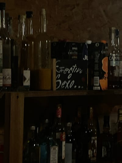 The image shows a shelf stocked with various bottles and containers, likely alcohol or mixers. Labels like "Campari", "Unifre", and "Festive Sexo..." are partially visible. The lighting is dim, and the image is somewhat blurry. Based on the labels and the context, it's likely a bar or restaurant in Puerto Escondido, Mexico. There is no presence of people in this image.