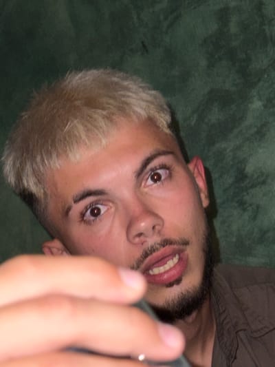 The image presents a close-up portrait of a young adult male, facing directly towards the camera. He has short, bleached blonde hair with darker roots, prominent dark eyebrows, one featuring a distinct shaved slit. His brown eyes are wide open, displaying a strong red-eye effect from direct flash illumination. A dark mustache and beard frame his mouth, which is slightly open to reveal a glimpse of his upper teeth. His complexion is fair, with some redness visible on his skin and ear. He is wearing a dark, possibly olive-green, collared shirt, visible on his right side (viewer's left). A blurred hand, likely his own, occupies the lower left foreground, extending towards the viewer and partially obscuring the lower portion of his face and body. The background is a uniformly textured, dark green surface, suggesting an indoor setting.