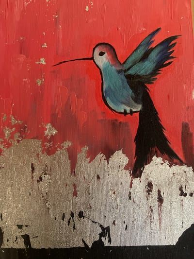 The image is a close-up of an acrylic painting on canvas. The painting depicts a hummingbird in flight against a red background. The hummingbird is rendered in shades of blue and red, with black accents for its tail and wingtips. There are flecks of what appear to be silver leaf scattered across the red background and in the lower portion of the painting, which also features a textured silver and black abstract element. The texture of the canvas is visible throughout the image. The painting is not located in the city of Shcherbinka, Russia; it is an artwork.