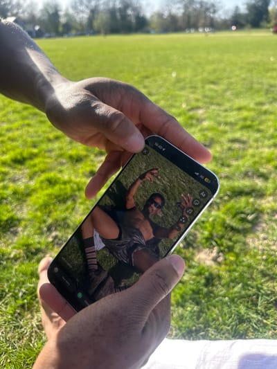 The image shows a person holding a smartphone outdoors, with the phone displaying a photograph of a man in sunglasses making a pose on a grassy field. The background of the scene is a blurred expanse of green grass, suggesting a park or open space. The photo on the phone appears to have been taken in a similar outdoor setting. The setting is possibly in the area of Tunbridge Wells, United Kingdom, implied by the landscape and atmosphere.