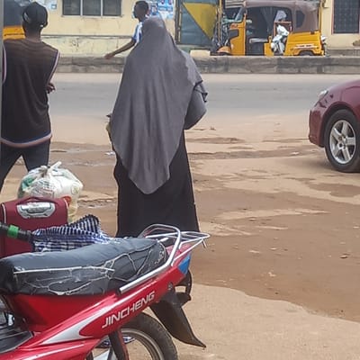 The image, taken in Sardauna, Nigeria, depicts a street scene. A red JinCheng motorcycle is parked on the right side of the frame, with items strapped to its back. Two individuals are visible: one wearing a black cap and a brown striped shirt, and another woman wearing a gray covering. A car is partially visible in the top of the frame, and yellow tricycles are visible on the side of the road. The environment appears to be a dusty, urban road with buildings lining the street.