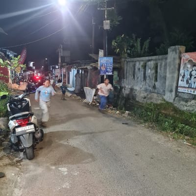 A young man wearing a blue shirt and dark pants is running down a street in Kecamatan Tamansari, Indonesia, at night. He is heading toward a group of people, likely a family, standing around a scooter. The man is running in the opposite direction of a woman in a light-colored shirt and dark pants, who is also running down the street but is heading toward a building on the right side of the street. The man and woman are both running on the right side of the street, which is made of asphalt. On the left side of the street is a sidewalk and a line of houses. There is a scooter parked on the right side of the street. The scene is lit by a streetlight.  The street is surrounded by trees. There is a sign on the side of one building and another sign that appears to be advertising a local business.