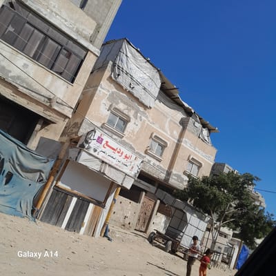 In Deir el-Balah, Palestinian Territories, the scene depicts a street with several buildings under construction or in a state of disrepair. The foreground is dominated by a sandy, uneven ground. To the left, a building shows a damaged window on its upper floor, with blue tarp material hanging from its lower section. Adjacent to this, a shop front is visible, featuring a sign with Arabic text. Further to the right, another building has a similar sign, also in Arabic, with a black and white image of a man. A tree with green foliage stands to the right of this building. In front of the buildings, two children, a boy and a girl, are walking on the sandy ground. The boy, wearing a white t-shirt and dark pants, is looking towards the girl. The girl, in an orange shirt and pants, is holding a green object. Behind the children, a trailer with a covered structure is parked. The sky is clear and blue. The overall impression is one of an active, but possibly impoverished, urban environment.