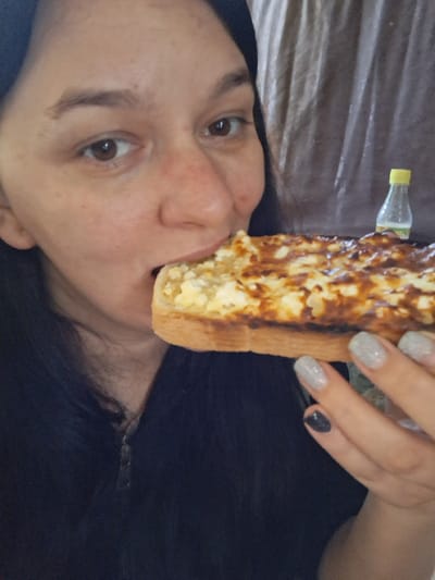 A woman is shown in a close-up shot, holding a slice of toasted bread with a topping of melted cheese and brown, crispy bits. She is taking a bite of the bread. Her nails are painted with a glittery silver polish on some fingers and dark grey on one. In the background, a small, clear bottle with a yellow cap is visible. The environment appears to be indoors, with a textured, draped fabric in the background. The image was taken in Marchevo, Bulgaria.