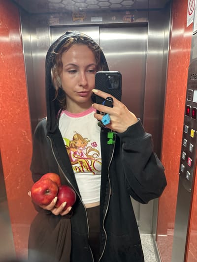 A young woman with curly brown hair is taking a selfie in an elevator. She is wearing a black zip-up hoodie over a white t-shirt with a cartoon character printed on it, and several rings on her fingers. She is holding a smartphone in her right hand and two red apples in her left. The elevator has orange marble-patterned walls and stainless steel doors. The elevator control panel is visible at the top of the image with several buttons and a red emergency button. The image is oriented vertically and appears to be taken in natural light. The overall mood is casual and playful. There is some visible text on the elevator control panel which appears to be in Cyrillic script.