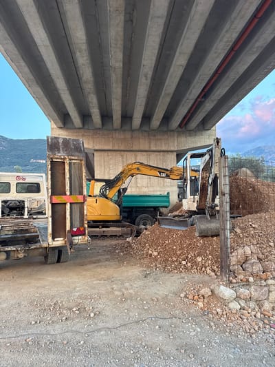 Under a concrete overpass in Burtaiši, Montenegro, construction equipment is present. A yellow CAT excavator is positioned in the middle ground, with its arm extended over a green dump truck. To the right of the CAT excavator, a smaller white Terex excavator is partially visible, its bucket resting in a pile of dirt. In the foreground, a large trailer with a weathered metal ramp is parked. Behind the trailer, a white truck is partially visible, with a speed limit sign showing "70 80". The ground is covered in dirt and gravel. The sky above is a clear blue with some clouds visible on the horizon. Mountains are visible in the background.