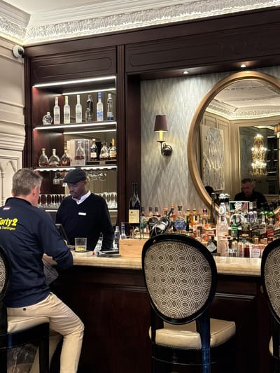 The image shows a bar scene. A bartender in a black sweater and cap is behind the bar, potentially serving a drink to a customer seated at the bar. The customer is wearing a navy jacket. Shelves filled with liquor bottles are visible behind the bar, with a mirror above reflecting the bar area and a partial view of what appears to be a person. The setting suggests a sophisticated establishment. While the city of Cape Town, South Africa, is mentioned, it is not verifiable as being there from only the scene in the picture.