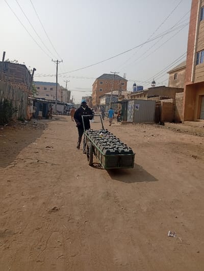 The image displays a dusty, unpaved road in Sabon Gari East, Nigeria, under a bright, hazy sky. A person, appearing male, dressed in dark attire and a light blue cap, is pushing a two-wheeled, green metal cart loaded with approximately twenty-five dark-colored rectangular plastic jerrycans. The road surface is reddish-brown dirt, exhibiting tire tracks and footprints, with a small piece of white litter visible in the lower right foreground. To the left, a series of makeshift structures constructed from corrugated metal sheets and wood line the street. On the right, more substantial multi-story buildings, predominantly light brown, are visible further down the road, interspersed with smaller concrete structures. In the distant background, several individuals are walking along the street. Overhead, multiple utility lines are strung across the scene, supported by wooden poles. A building in the far background on the right possesses two small, distinct domed features on its roof.