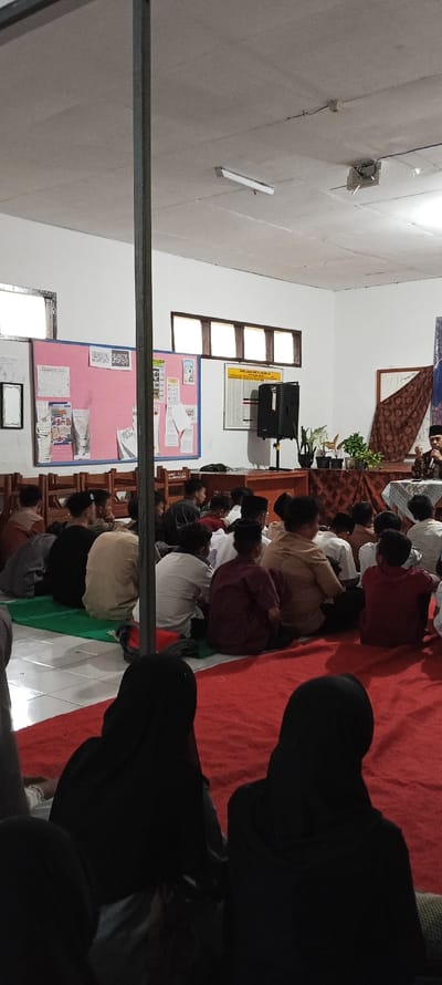 The image shows a gathering of people inside a building in Cangkuang, Indonesia (latitude: -7.065, longitude: 107.552). The room has a white ceiling with a single fluorescent light fixture and a white tiled floor. The walls are painted white. There are three windows with wooden frames along the back wall. A large bulletin board covered in pink paper with white lettering and multiple pieces of paper is on the left wall. A wooden desk with brown paper covering it is on the right side of the image. There is a large speaker on top of the desk. A black curtain with a pattern of white and gold squares is draped over the right side of the desk. A small, potted plant sits on the desk in front of the curtain. Another potted plant is on a shelf behind the desk. There are several rows of wooden chairs lining the side walls. There are 20 people sitting on the floor, most of whom are wearing traditional Muslim clothing. There are two women in the foreground of the image, wearing black hijabs that fully cover their heads and faces. The people are looking in the direction of an individual sitting on a chair in front of the curtain. The individual in the chair is wearing a white shirt, a white skullcap, and a black jacket. They are holding a black microphone. The people on the floor appear to be listening to the speaker.