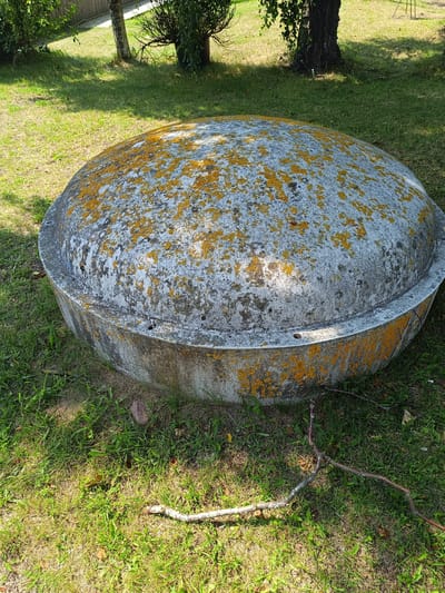 A domed, circular concrete structure covered in patches of orange lichen sits on a patch of grass in Pāvilosta, Latvia. The structure is low to the ground with a thick, reinforced rim. The surrounding area is grassy with scattered leaves and a few fallen twigs. In the background, trees and a wooden fence are visible, suggesting an outdoor, possibly residential or park-like setting. The sun casts shadows on the grass, indicating it is daytime. There are no people visible in the image.