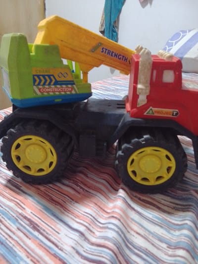 A toy crane truck is displayed on a bed with striped sheets. The truck is red and green with yellow accents, featuring a crane arm that extends from the top. The crane arm has the word "STRENGTH" printed on it in blue letters. The truck's body is labeled "CRANE CONSTRUCTION" in blue and red lettering. The truck has large black wheels with yellow rims. A sticker with a green triangle and the word "PROJECT" in red is visible on the side of the red cab. The background is blurred, showing a white wall and a portion of a patterned pillow. The setting appears to be indoors, possibly a bedroom, given the presence of the bed and wall. No people are visible in the image.
