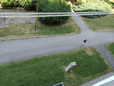 The image is a high-angle aerial view of an outdoor scene in Košice, Slovakia, likely a residential area or park. A single person, appearing to be a young adult wearing a dark red shirt, white shorts, and carrying a light-colored bag over their shoulder, is walking on a paved path. The path winds through areas of green grass and patches of dry, yellowed grass.
Surrounding the path are dense green bushes and trees, with some larger trees casting shadows. Two yellow poles with black bands are visible, one on the left and one in the middle ground, possibly streetlights or utility poles. In the lower right quadrant, a light-colored concrete structure is partially visible, possibly a bollard or marker. In the far foreground, a railing with a light blue accent is visible, suggesting this is taken from a balcony or elevated structure. Two thin, horizontal wires stretch across the upper portion of the image. The overall environment appears to be a well-maintained outdoor public space.