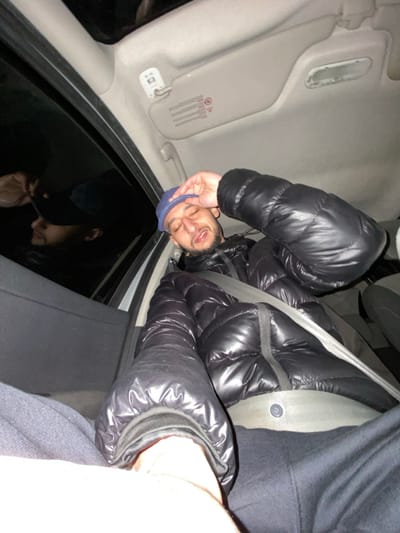 A man in a black puffer jacket and blue baseball cap is upside down in a car. His face is visible, and he is secured with a seatbelt. The car's interior includes a sun visor and window. The dark window reflects the man's face. Part of another person's clothing is visible on the right side.