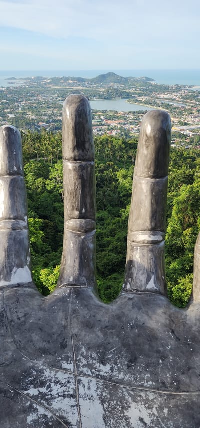 The image shows a close-up view of concrete fingers from a statue, likely a giant hand, overlooking a lush, tropical landscape and a coastal town. The statue appears weathered, with patches of gray and white on its surface, indicating exposure to the elements. The fingers are segmented, with clear lines where each section joins.

In the background, a dense canopy of green trees, characteristic of a tropical rainforest, covers the hills. Beyond the foliage, a town with colorful rooftops stretches towards a body of water, possibly the sea or a large bay. The water is a calm, pale blue, meeting a hazy horizon under a bright sky. The overall scene suggests a sunny day.

The perspective is from high up, looking down and outwards, implying the statue is situated on a hilltop or a high vantage point. The orientation of the image is vertical, with the fingers extending downwards. The presence of the statue's hand suggests a tourist attraction or a religious monument, common in locations like Ko Samui, Thailand, known for its natural beauty and spiritual sites. No people are visible in the frame, and there is no visible text. The focus is on the texture and scale of the statue in contrast with the vast natural and urban landscape below.