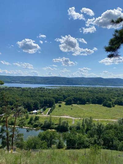 An elevated, wide-angle shot captures a panoramic view of the Mastryukovo region in Russia on a bright, sunny day with scattered white clouds against a clear blue sky. The foreground is dominated by a grassy slope with some sparse vegetation and a glimpse of wooden railings. Below, a winding river or lake is visible, bordered by lush green trees and a small pier. Further back, a dense forest stretches across the landscape, interspersed with open green fields. A dirt road meanders through the fields, and a small cluster of buildings, including a white tent-like structure, can be seen among the trees. In the distance, a wide body of water, likely the Volga River, is prominent, with a range of low, tree-covered hills forming the horizon line. A faint scattering of buildings is visible on the distant hills. A single pine branch with needles is in the upper right corner of the frame.