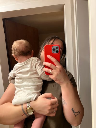 This image captures an adult holding an infant, depicted as a mirror selfie taken indoors within Wüstenrot, Germany. The adult, possessing dark hair, has her face partially obscured by a red-cased smartphone held in her right hand, actively capturing the reflection. She wears a dark olive-green ribbed t-shirt. A silver band ring is present on her right ring finger, and multiple thinner rings are worn on her left hand. Visible tattoos include an abstract design on the upper left arm and a distinct shoe or boot outline on the lower right arm. A gold-toned rectangular watch with a metal band and a red string bracelet are worn on her left wrist. Minor reddish marks are visible on the dorsal surface of her right hand. The infant, held facing away from the viewer against the adult's body, displays the back of its head with light-colored, fine curly hair exhibiting some thinning at the crown. The infant is clad in a white long-sleeved onesie patterned with subtle light-colored designs. The background, seen as a reflection in the mirror, includes a light-colored doorframe and a brown wooden door.