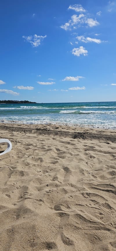 A wide-angle shot captures a serene beach scene in Primorsko, Bulgaria. The foreground is dominated by textured sand, showing footprints and small pebbles. A portion of a white object, possibly a beach chair, is visible on the left side. The middle ground features gentle waves lapping onto the shore, creating white foam. Beyond the water, a distant coastline with buildings and greenery stretches across the horizon. The sky is a vibrant blue, dotted with scattered white clouds. The overall impression is one of a calm, sunny day at the seaside.