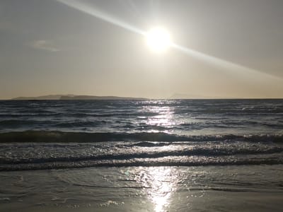 The image depicts a sunlit seascape, likely captured in the coastal area of Agios Stefanos, Greece. The sun is positioned high in the sky, casting a bright glare and creating a prominent reflection on the water's surface. The sea is moderately choppy, with small waves visible closer to the shore. The water reflects the sunlight, creating a shimmering effect. In the distance, a faint outline of land is visible, likely a neighboring island or a distant part of the coast. The sky appears hazy or overcast, contributing to the overall brightness of the scene. No people or distinct objects are visible in the image apart from the natural elements. The overall atmosphere suggests either early morning or late afternoon light conditions.