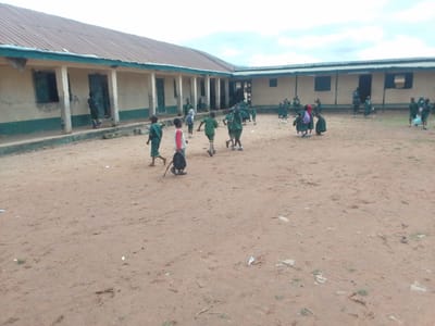 The image depicts an outdoor scene at a school in Balewa, Nigeria, with a group of young children, likely students, in the foreground and midground. The children are predominantly wearing green uniforms. The school building, characterized by its light-colored walls, a corrugated metal roof, and an open walkway with pillars, forms the backdrop. The ground is unpaved, consisting of sandy dirt with some scattered debris. Several children are engaged in walking and running activities, suggesting a moment of recreation or transition between activities. The sky is overcast, indicating a cloudy day.