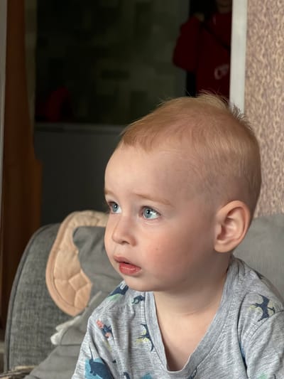 The image is a close-up, profile view of a young child with light blonde hair and blue eyes. The child is wearing a grey t-shirt with a pattern of what appear to be stylized dinosaurs. The child is looking to the left of the frame with an attentive expression. The background is blurred, but suggests an indoor setting with a textured wall to the right and a dark area to the left that might be a doorway or window. A portion of a grey sofa with a light-colored pillow is visible on the left side of the frame. The image does not contain enough information to determine a specific location in the city of Уральское, Russia.