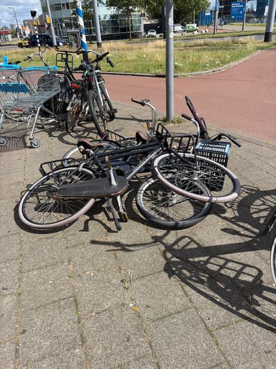 This is a ground-level, eye-level, outdoor, daylight photograph taken in the city of Rotterdam, Netherlands. The foreground is dominated by several bicycles that appear to have been haphazardly left or dropped on a paved sidewalk. One bicycle is lying on its side, with its wheels and frame spread out. Another is leaning against a pole, and parts of others are visible, also on the ground. A metal shopping cart with blue plastic accents is positioned to the left of the bicycles. The sidewalk is made of grey concrete paving stones. In the mid-ground, a narrow strip of dry grass and weeds separates the sidewalk from a red asphalt path. Beyond the path, there is a grassy area and a metal fence. In the background, several buildings can be seen, including a modern building with glass windows. A street light and utility pole are also present. The sky is partly cloudy. There are no people visible in the image. The overall impression is one of disarray and neglect of the bicycles and shopping cart.