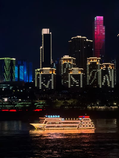 The image displays a detailed night scene of the Nan'an District, China, featuring a brightly illuminated passenger boat on a body of water, likely a river, against a backdrop of a vibrant city skyline.
In the immediate foreground, dark water shows subtle ripples and reflections of the boat's lights.
The primary subject is a multi-decked passenger boat, approximately centrally located, moving from left to right. The boat is extensively outlined and adorned with warm yellow-orange lights. Its stern features several large, arched window-like structures. On the upper deck, two sets of Chinese characters are illuminated: "朝天门" (Cháotiānmén) in blue on the left and "两江游" (Liǎngjiāng Yóu) in red on the right. No discernible individuals are visible on board.
The background reveals a densely built city skyline. On the far right, a tall skyscraper is distinctly illuminated in a vivid magenta or pink color across its facade. To its left, several dark residential or commercial buildings feature a pattern of white internal window lights. Further left, a cluster of buildings showcases detailed yellow-orange decorative lighting on their rooftops and upper sections, with internal window lights forming geometric patterns, some resembling abstract letters like "M". In the central-left area, two prominent, slender skyscrapers are illuminated with vertical lines of yellow-white light. Towards the far left, a group of buildings is illuminated with dynamic vertical strip lighting in green and blue hues. Below the main skyline, a strip of land along the riverbank is dotted with numerous smaller, varied lights, including some red accents. The sky above is dark, indicating a clear night.