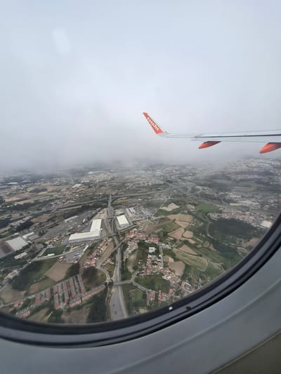The image shows a view from an EasyJet airplane window, looking down over Matosinhos, Portugal. The wing of the plane is visible in the upper left, with an orange "easyJet" flag attached. The view below reveals a landscape comprised of a mix of urban and rural areas. There are densely populated residential areas, industrial zones with large warehouse-type buildings, fields, and roads including highway overpasses. The weather appears overcast, with some areas obscured by fog or low clouds.