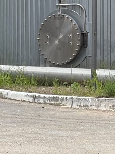 The image shows a large, round, metal hatch with several bolts around its circumference, affixed to a corrugated metal wall. A metal pipe runs horizontally in front of the wall, partially obscured by grass. The ground in the foreground is paved with asphalt, bordered by a concrete curb. The scene appears to be outdoors, possibly in an industrial or utility area. There are no people visible in the image. The location is identified as Syktyvkar, Russia.