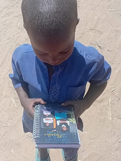 A high-angle shot depicts a young child, appearing to be male, standing outdoors on a light brown, unpaved sandy surface with scattered dry vegetation in Ayama, Nigeria. The child wears a blue short-sleeved top with white embroidered patterns on the chest and sleeves, light-colored trousers, and light-colored footwear. The child holds a spiral-bound notebook with both hands, looking down at its cover. The notebook cover is dark, featuring four small portrait photographs arranged in a grid, with visible text including "Thanks" in a decorative font and "J. Godwin." The child's head is tilted forward, obscuring most of their face as they focus on the notebook. The scene is illuminated by natural daylight.