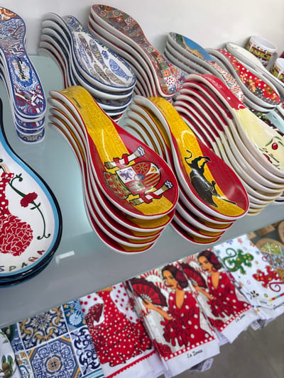 The image shows a display of Spanish-themed ceramic spoon rests on shelves. The spoon rests are stacked and feature various designs, including the Spanish flag with a bull, floral patterns, and traditional Spanish imagery like a flamenco dancer. In the background, there are also stacks of plates and some textiles with similar Spanish motifs, such as the flamenco dancer and lizards. The scene suggests a souvenir shop or a market stall in Orihuela, Spain, selling traditional Spanish crafts and souvenirs. The overall lighting is bright, indicating an indoor setting, possibly a retail space. There are no people visible in the image.