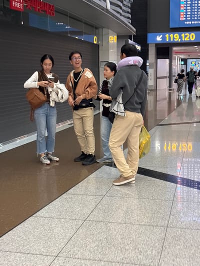 Incheon International Airport, South Korea, is depicted in this image. Four individuals are visible standing in an indoor concourse area. On the left, a woman with dark hair and glasses is holding a smartphone and a brown bag. Next to her, a person in a brown hooded jacket and tan pants is looking to their right. A third person, also with dark hair, is wearing a black and white striped shirt and stands in front of a dark wall with a gray roller shutter. On the right, a person in a gray sweatshirt and tan pants is wearing a purple neck pillow and holding a yellow plastic bag. In the background, a digital flight information board displays flight numbers and destinations. The floor is tiled with large gray and brown tiles.