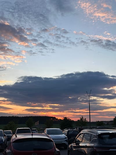 A wide-angle, eye-level shot captures a parking lot filled with cars at sunset in Бараўлянскі сельскі Савет, Belarus. The sky is dominated by dramatic clouds, with hues of orange, pink, and gray, indicative of the setting sun. A tall, dark lamp post stands prominently in the middle ground. In the background, a dark line of trees separates the parking lot from a field or more distant landscape, and a few buildings are visible on the far right. The foreground and mid-ground are occupied by several cars, their shapes silhouetted against the bright sky. The image appears to be taken from a slightly elevated position, looking down on the parked vehicles.