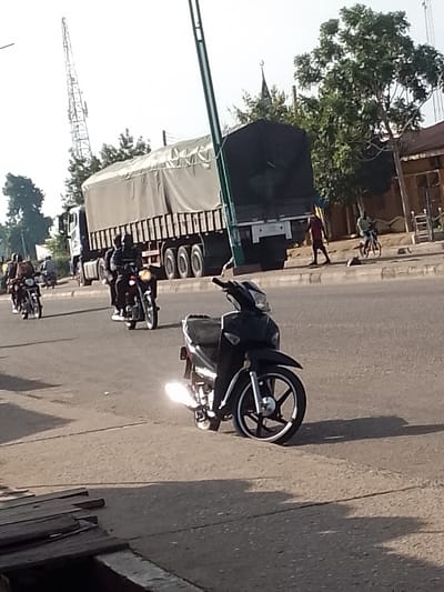 This is an eye-level, medium-shot, vertical photograph of a busy street scene, likely in Pantame, Nigeria, given the presence of a tall communication tower in the background, a common landmark in many Nigerian cities. The foreground is dominated by the rear of a black motorcycle, its bright headlight casting a strong glare. The middle ground shows a black motorcycle with a rider and a passenger, also illuminated by its headlight, moving away from the camera. Further back, a large, grey, covered truck is visible with multiple axles and wheels, suggesting it's a heavy-duty vehicle. Several other motorcycles, some with riders, are also present on the road, indicating active traffic. The environment appears to be an urban setting with buildings and trees lining the street on the right side. The ground is paved, and a sidewalk is visible. The lighting suggests it is daytime, likely with the sun at a low angle, casting shadows. The overall impression is one of a typical day in a bustling Nigerian city, with a focus on vehicular movement and activity.