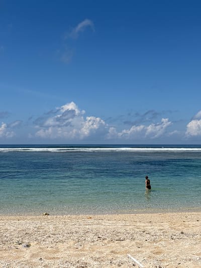 A person is standing waist-deep in the clear turquoise water of a beach. The shoreline is visible on the right, and it's covered with light-colored sand and small shells. The sky above is a bright blue with scattered white clouds, suggesting a sunny day. The location is likely Kutuh, Indonesia.